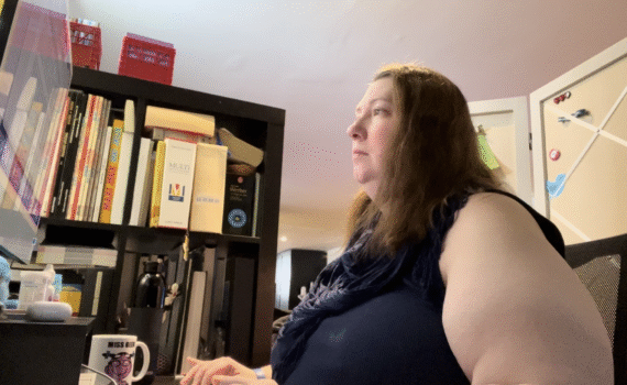 Annie-Danielle assise devant un écran d'ordinateur et qui tape sur un clavier, à côté d'une bibliothèque. On devine une tasse à café sur le bureau.